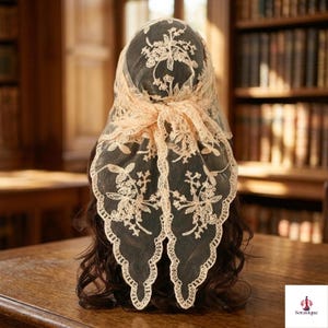 May include: A light peach-colored lace head covering featuring intricate floral embroidery and a scalloped edge. The sheer fabric is draped over dark hair, with a knotted detail at the back creating a triangular shape that hangs down.