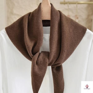 May include: A solid brown knit scarf draped around a wooden hanger over a white collared shirt. The scarf features a soft, textured fabric with a triangular front fold. The bottom right corner displays the text Soravique with a small logo.