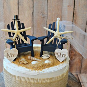 May include: A wedding cake topper featuring two miniature blue Adirondack chairs with starfish and heart-shaped signs that say "Mr." and "Mrs.". The chairs are decorated with a small top hat and a white veil.