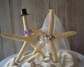 May include: A wedding cake topper featuring two starfish, one wearing a black top hat and a purple bow tie, and the other wearing a white veil and a floral crown. The starfish are standing on a bed of sand.