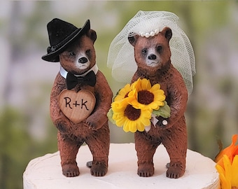 May include: A two-tiered wedding cake decorated with a rustic theme. The cake is adorned with sunflowers, roses, and greenery. A bride and groom bear cake topper is on top of the cake. The groom bear is wearing a black hat and a bow tie. The bride bear is wearing a white veil and holding a bouquet of sunflowers. The cake topper has a wooden heart with the initials "R+K" on it.