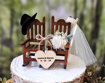 Könnte beinhalten: Rustikale Hochzeitstorte mit Birkenrinden-Optik, gekrönt von Miniatur-Schaukelstühlen aus Holz, einem schwarzen Hut und einem herzförmigen Anhänger mit der Aufschrift "Aaron & Stacey 4-21-23". Dekoriert mit Grün und weißen Blumen.