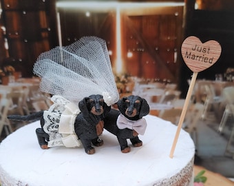 May include: A white wedding cake decorated with a floral garland and a topper of two dachshund figurines wearing a veil and a bow tie. The topper says "Just Married" on a heart-shaped sign.