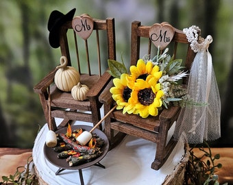 Op de afbeelding: Een bruidstaarttopper met twee houten schommelstoelen, een met een zwarte cowboyhoed en de andere met een witte sluier. De stoelen zijn versierd met harten waarop 'Mr.' en 'Mrs.' staat. Er is een klein kampvuur met marshmallows die roosteren voor de stoelen, en een zonnebloemenboeket op de stoel met de sluier.
