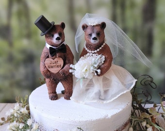 Könnte beinhalten: Eine weiße Hochzeitstorte mit einem braunen Bärenpaar als Dekoration. Der Bräutigam-Bär trägt einen schwarzen Zylinder und eine Fliege, während die Braut-Bärin einen weißen Schleier trägt und einen Strauß weißer Blumen hält. Die Torte ist mit einem Holzschild verziert, auf dem "Just Married" steht.