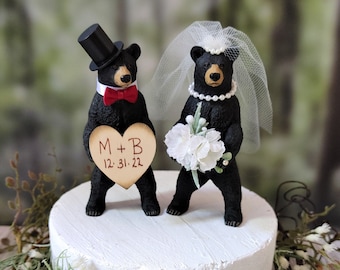 May include: Black bear wedding cake topper with a top hat and a veil. The groom bear holds a heart-shaped sign that says "M + B 12.31.22".