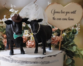 Op de afbeelding: Een witte cake met zwarte koeienfiguurtjes erop. De cake staat op een witte cake standaard. De cake is versierd met bloemen en een hartvormig bord met de tekst "Gonna love you until the cows come home."