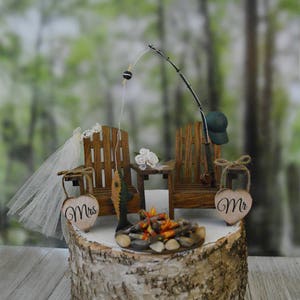 May include: A wedding cake topper featuring a miniature campfire, two wooden chairs, a fishing rod, and two hearts with the words "Mr." and "Mrs." on them.