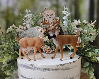Könnte beinhalten: Ein rustikaler Hochzeitskuchen, gekrönt von zwei Hirschfiguren, verziert mit Blumen und Grün. Der Kuchen hat ein weißes Frosting mit einem semi-nackten Design. Ein Holzschild liest "The hunt is over".
