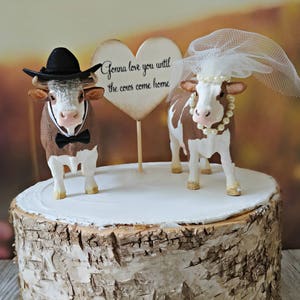 Op de afbeelding: Een bruidstaarttopper met twee bruine en witte koeien, een met een zwarte cowboyhoed en een bruine strik, en de andere met een witte sluier. De topper bevat een hartvormig bord met de tekst "Gonna love you until the cows come home."