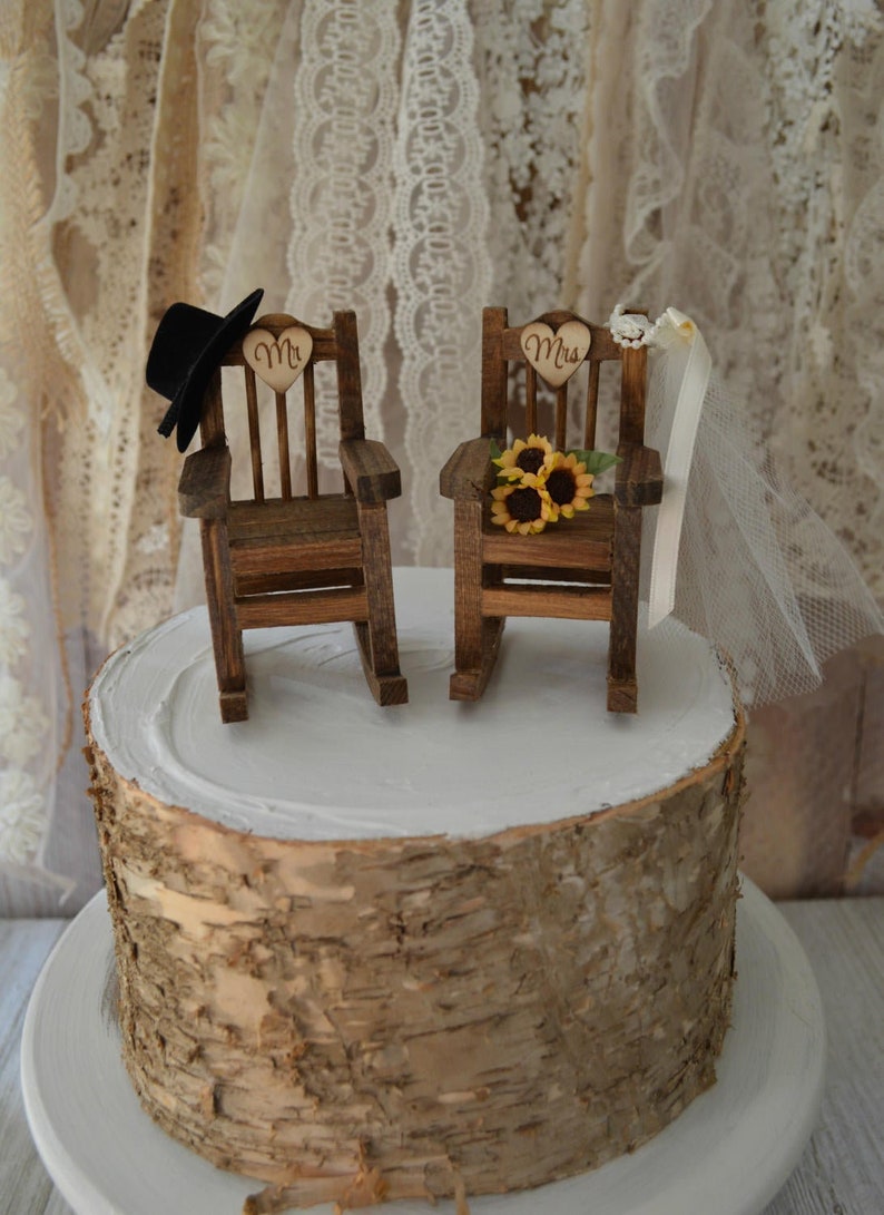 Puede incluir: Un adorno de pastel de boda r&uacute;stico con dos mecedoras de madera, una con un sombrero de copa negro y la otra con un velo blanco y girasoles. Las sillas est&aacute;n decoradas con letreros en forma de coraz&oacute;n que dicen "Mr." y "Mrs."