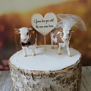 Op de afbeelding: Een bruidstaarttopper met een bruine en witte koe met een strikje en een bruine en witte koe met een sluier en een parelketting. De topper staat op een houten taartstandaard en bevat een hartvormig bord met de tekst "Gonna love you until the cows come home."