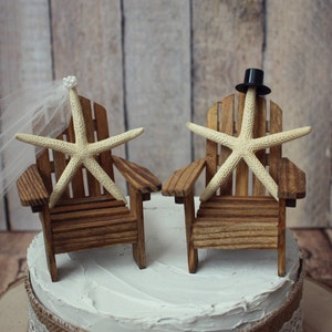 May include: A wedding cake topper featuring two miniature wooden chairs with starfish sitting on them. The bride's chair has a white veil and the groom's chair has a black top hat.