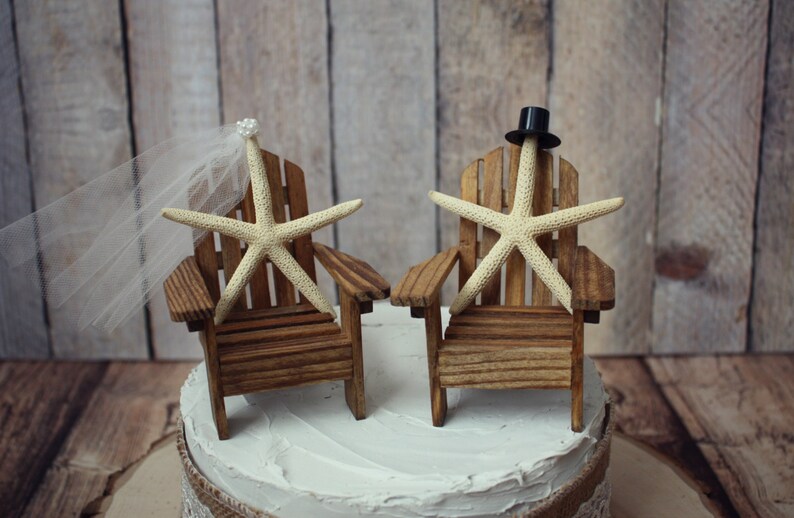 May include: A wedding cake topper featuring two miniature wooden chairs with starfish sitting on them. The bride's chair has a white veil and the groom's chair has a black top hat.