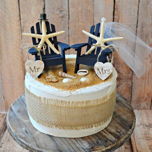 May include: A white wedding cake decorated with a burlap ribbon and a beach-themed topper. The topper features two blue Adirondack chairs with starfish and a veil. The chairs are decorated with wooden hearts that say "Mr." and "Mrs."