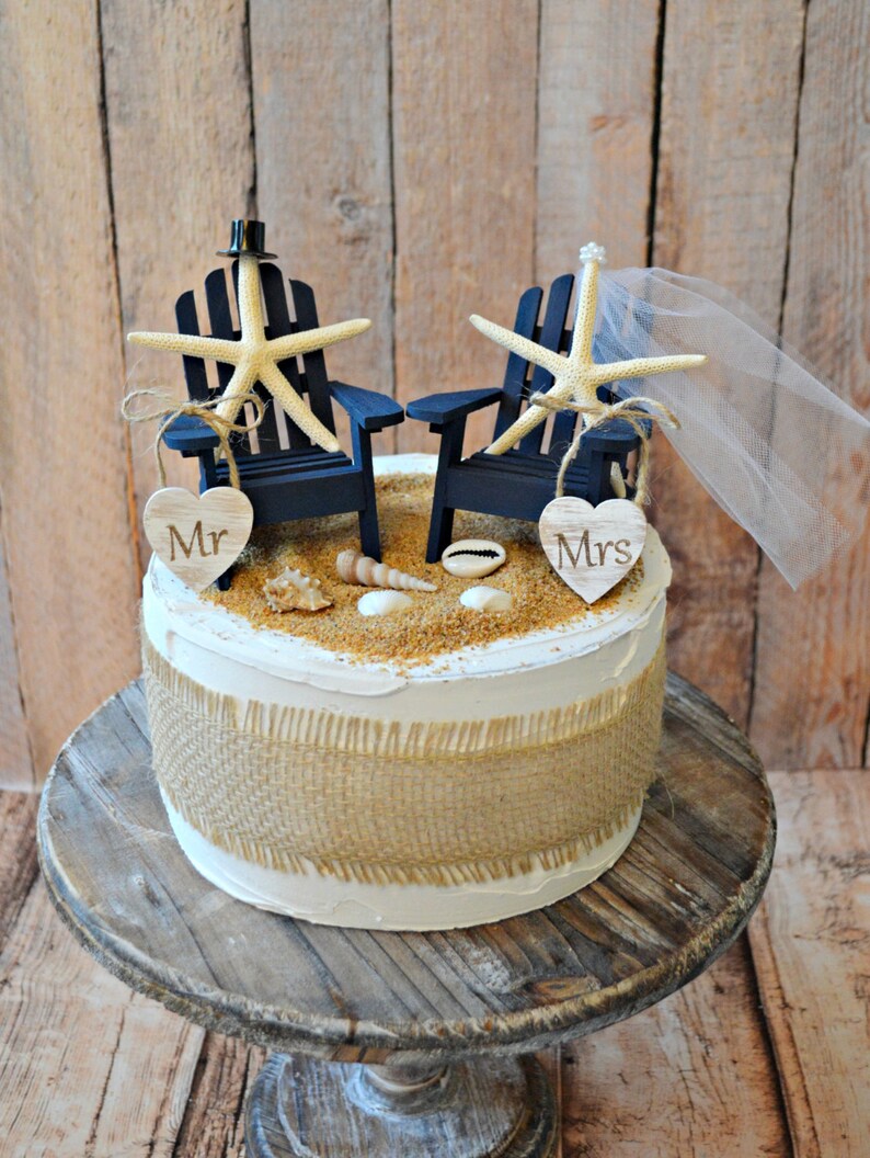 May include: A white wedding cake decorated with a burlap ribbon and a beach-themed topper. The topper features two blue Adirondack chairs with starfish and a veil. The chairs are decorated with wooden hearts that say "Mr." and "Mrs."