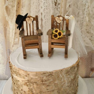 Puede incluir: Un adorno de pastel de boda r&uacute;stico con dos mecedoras de madera, una con un sombrero de copa negro y la palabra "Mr." en un coraz&oacute;n, y la otra con un velo blanco y la palabra "Mrs." en un coraz&oacute;n. Las sillas est&aacute;n decoradas con girasoles.
