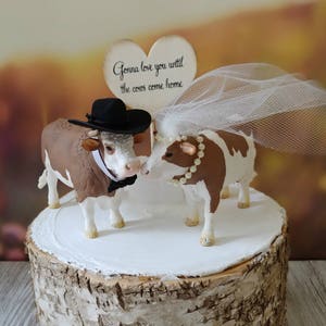 Op de afbeelding: Een bruidstaarttopper met een bruine en witte koe die een zwarte hoed en een strikje draagt, en een bruine en witte koe die een witte sluier draagt. De topper staat op een houten taartstandaard en bevat een bord met de tekst "Gonna love you until the cows come home."