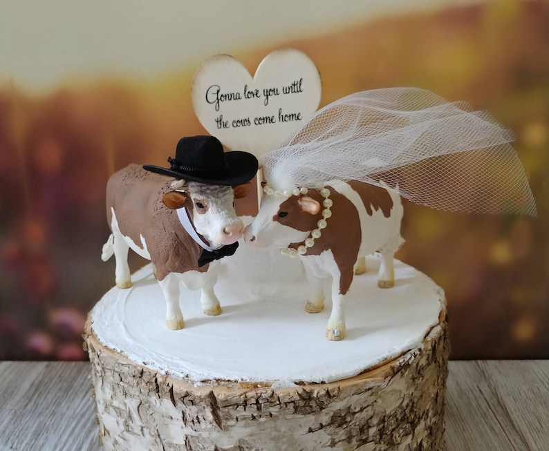 Op de afbeelding: Een bruidstaarttopper met een bruine en witte koe die een zwarte hoed en een strikje draagt, en een bruine en witte koe die een witte sluier draagt. De topper staat op een houten taartstandaard en bevat een bord met de tekst "Gonna love you until the cows come home."