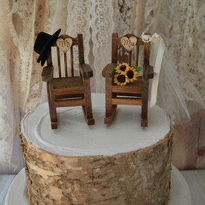Puede incluir: Un adorno de pastel de boda r&uacute;stico con dos mecedoras de madera, una con un sombrero de copa negro y la otra con un velo blanco. Las sillas est&aacute;n decoradas con letreros en forma de coraz&oacute;n que dicen "Mr." y "Mrs." y un ramo de girasoles.