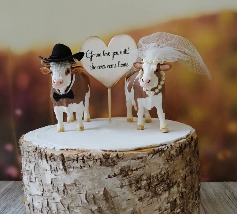 Op de afbeelding: Een bruidstaarttopper met een bruine en witte koe die een zwarte hoed draagt en een bruine en witte koe die een witte sluier draagt. De topper bevat een hartvormig bord met de tekst "Gonna love you until the cows come home."