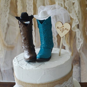 May include: A two-tiered wedding cake decorated with burlap and lace. The cake topper features a pair of cowboy boots, one brown and one teal blue, with a cowboy hat and a veil. A wooden heart with the text "Mr. & Mrs." is also included.