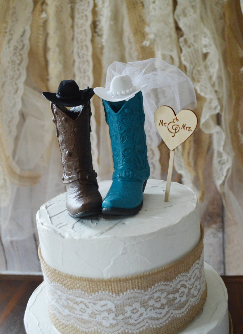 May include: A two-tiered wedding cake decorated with burlap and lace. The cake topper features a pair of cowboy boots, one brown and one teal blue, with a cowboy hat and a veil. A wooden heart with the text "Mr. & Mrs." is also included.