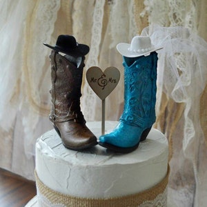 May include: A wedding cake topper featuring a pair of cowboy boots, one brown and one blue, with hats on top. The boots are decorated with floral designs. The bride's boot has a white veil. The topper includes a small heart-shaped sign that says "Mr. & Mrs."