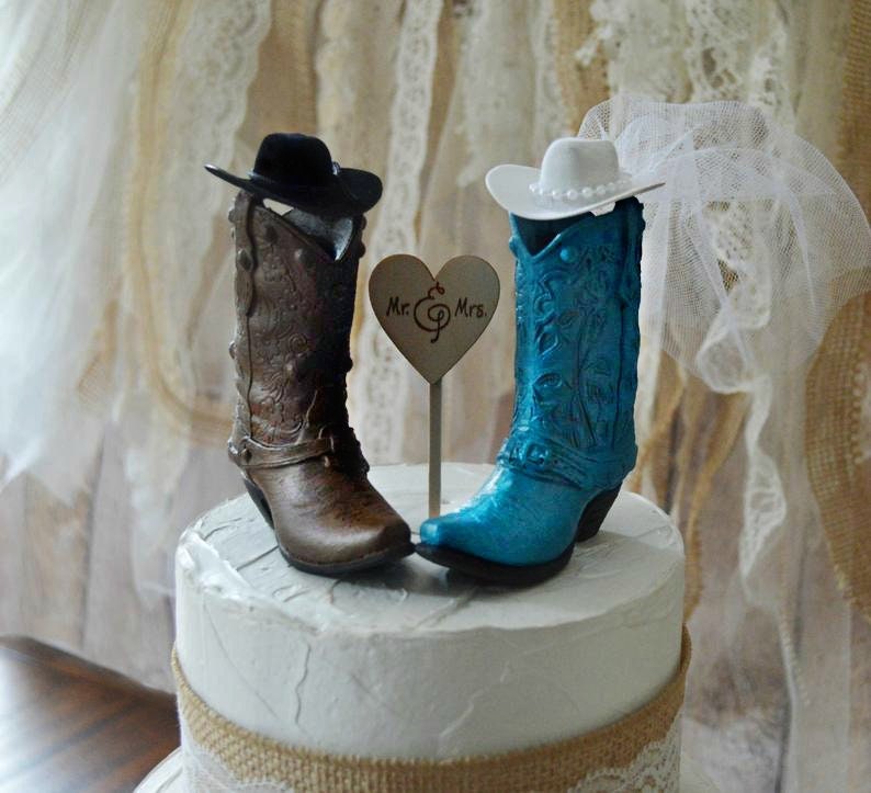 May include: A wedding cake topper featuring a pair of cowboy boots, one brown and one blue, with hats on top. The boots are decorated with floral designs. The bride's boot has a white veil. The topper includes a small heart-shaped sign that says "Mr. & Mrs."