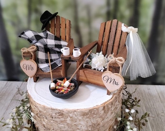 May include: A wedding cake topper featuring two wooden chairs, a black and white checkered blanket, a small fire pit with marshmallows, and two wooden hearts that say "Mr." and "Mrs."