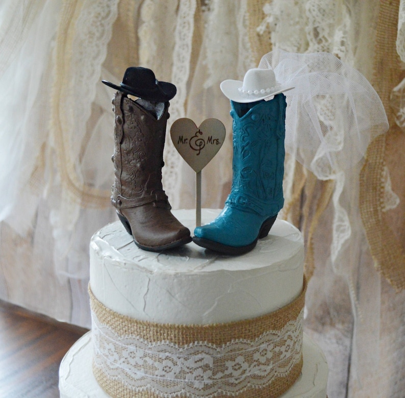 May include: A wedding cake topper featuring a pair of cowboy boots, one brown and one turquoise blue, with hats and a heart-shaped sign that reads "Mr. & Mrs."