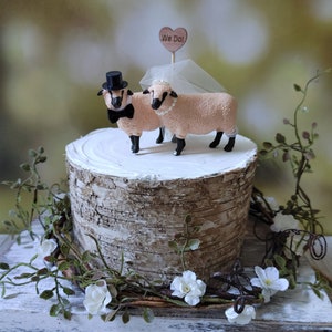 Pu&ograve; includere: Un topper per torta nuziale con una statuina di pecora sposa e sposo. Le pecore indossano un cappello a cilindro nero e un velo bianco. Il topper per torta &egrave; su un supporto per torta in corteccia di betulla con un cartello a forma di cuore in legno che dice "We Do!"