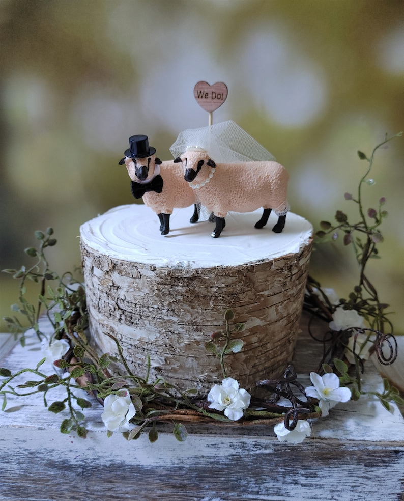 Pu&ograve; includere: Un topper per torta nuziale con una statuina di pecora sposa e sposo. Le pecore indossano un cappello a cilindro nero e un velo bianco. Il topper per torta &egrave; su un supporto per torta in corteccia di betulla con un cartello a forma di cuore in legno che dice "We Do!"