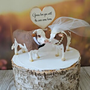 Op de afbeelding: Een bruidstaarttopper met een bruine en witte stier en een bruine en witte koe met een witte sluier. De topper staat op een taartstandaard van berkenhout. Het bordje zegt "Gonna love you until the cows come home."