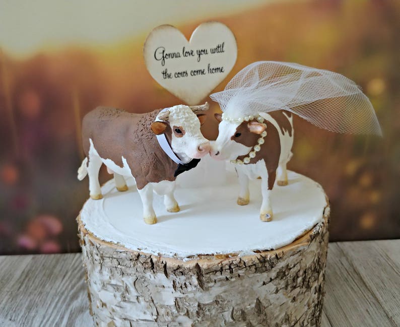 Op de afbeelding: Een bruidstaarttopper met een bruine en witte stier en een bruine en witte koe met een witte sluier. De topper staat op een taartstandaard van berkenhout. Het bordje zegt "Gonna love you until the cows come home."