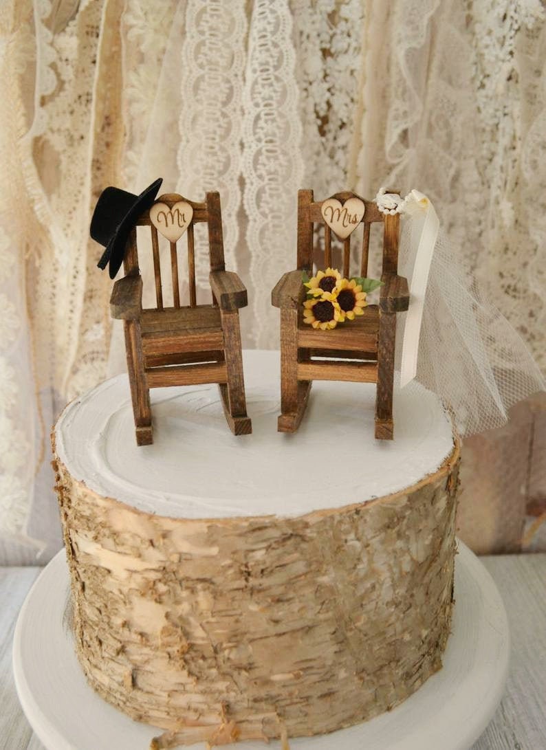 Puede incluir: Un adorno de pastel de boda r&uacute;stico con dos mecedoras de madera, una con un sombrero de copa negro y la otra con un velo y girasoles. Las sillas est&aacute;n decoradas con letreros en forma de coraz&oacute;n que dicen "Mr." y "Mrs."