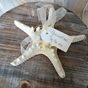May include: A cream-colored starfish with a tag that reads "You and me by the sea" tied with a sheer ribbon. The starfish is resting on a weathered wooden surface, evoking a coastal theme.