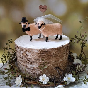 Pu&ograve; includere: Un topper per torta nuziale con due figurine di pecore, una con un cappello a cilindro e un papillon, l'altra con un velo. Il topper &egrave; posizionato su un supporto per torta in corteccia di betulla e include un piccolo cartello di legno a forma di cuore che dice "We Do!"