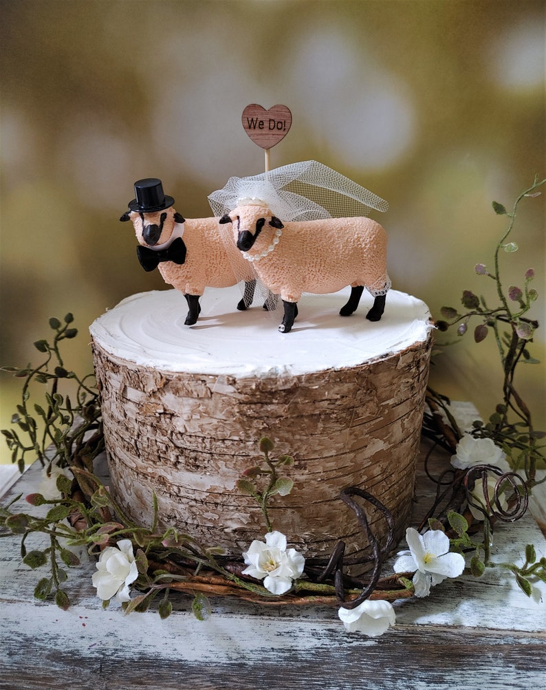 Pu&ograve; includere: Un topper per torta nuziale con due figurine di pecore, una con un cappello a cilindro e un papillon, l'altra con un velo. Il topper &egrave; posizionato su un supporto per torta in corteccia di betulla e include un piccolo cartello di legno a forma di cuore che dice "We Do!"