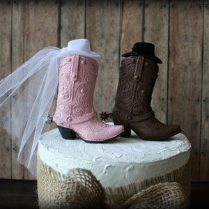 May include: A pink and brown cowboy boot cake topper with a white veil on the pink boot. The boots are on a white cake with a burlap base.