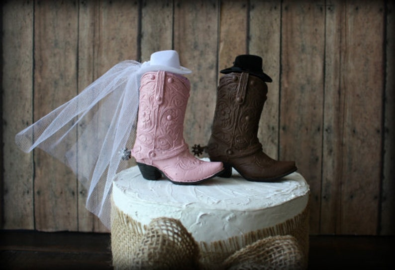 May include: A pink and brown cowboy boot cake topper with a white veil on the pink boot. The boots are on a white cake with a burlap base.