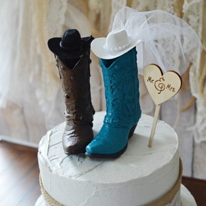 May include: A three-tiered wedding cake decorated with burlap and lace. The top tier features a cowboy boot cake topper with a brown boot for the groom and a teal boot for the bride. The bride's boot has a white cowboy hat and a white veil. The cake topper also includes a wooden heart with the text "Mr. & Mrs."