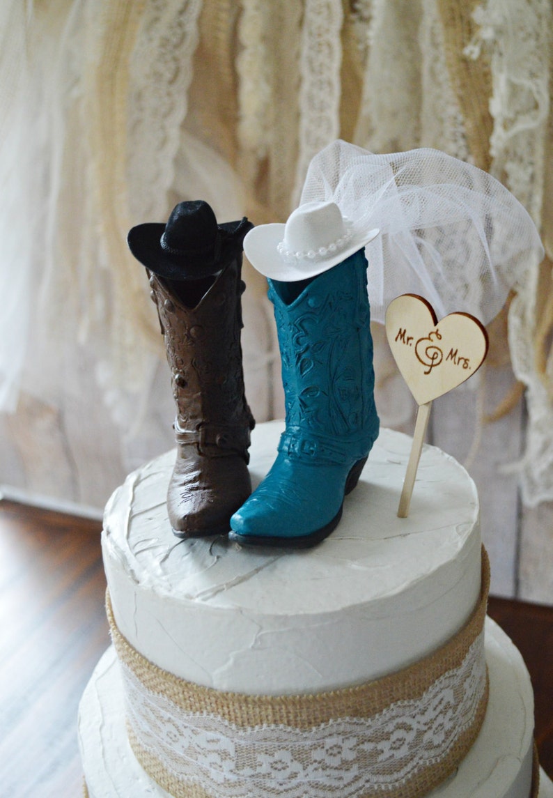May include: A three-tiered wedding cake decorated with burlap and lace. The top tier features a cowboy boot cake topper with a brown boot for the groom and a teal boot for the bride. The bride's boot has a white cowboy hat and a white veil. The cake topper also includes a wooden heart with the text "Mr. & Mrs."