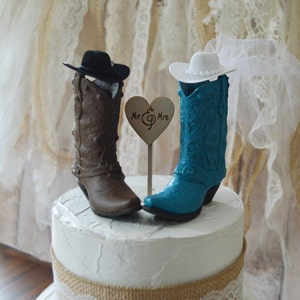 May include: A wedding cake topper featuring a pair of cowboy boots, one brown and one teal blue. The boots are wearing hats, the teal boot has a white veil. The topper includes a wooden heart with the text "Mr. & Mrs."