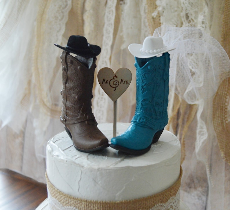 May include: A wedding cake topper featuring a pair of cowboy boots, one brown and one teal blue. The boots are wearing hats, the teal boot has a white veil. The topper includes a wooden heart with the text "Mr. & Mrs."