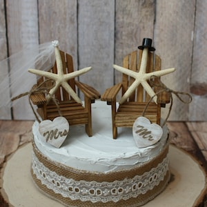 May include: A wedding cake topper featuring two miniature wooden chairs with starfish and a bride and groom topper. The bride has a veil and the groom has a top hat. The chairs are decorated with burlap and lace. The cake topper is on a white cake with a burlap and lace ribbon.