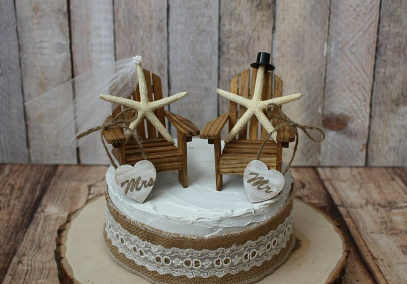 May include: A wedding cake topper featuring two miniature wooden chairs with starfish and a bride and groom topper. The bride has a veil and the groom has a top hat. The chairs are decorated with burlap and lace. The cake topper is on a white cake with a burlap and lace ribbon.