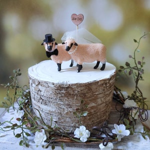 Pu&ograve; includere: Un topper per torta nuziale con due figurine di pecore, una con un cappello a cilindro e un papillon, l'altra con un velo. Il topper &egrave; posizionato su un supporto per torta in corteccia di betulla con un cartello a forma di cuore in legno che dice "We Do!"