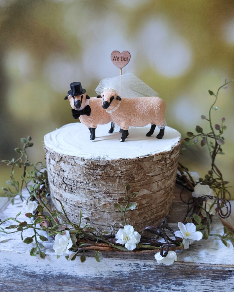 Pu&ograve; includere: Un topper per torta nuziale con due figurine di pecore, una con un cappello a cilindro e un papillon, l'altra con un velo. Il topper &egrave; posizionato su un supporto per torta in corteccia di betulla con un cartello a forma di cuore in legno che dice "We Do!"