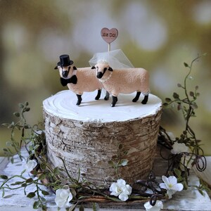Pu&ograve; includere: Un topper per torta nuziale con due figurine di pecore, una con un cappello a cilindro e un papillon, l'altra con un velo. Il topper &egrave; posizionato su un supporto per torta in corteccia di betulla con un cartello a forma di cuore in legno che dice "We Do!"